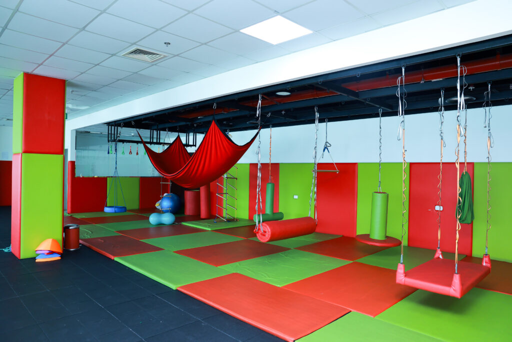 Colorful sensory gym room equipped with swings, therapy balls, and padded flooring for children’s developmental therapy.