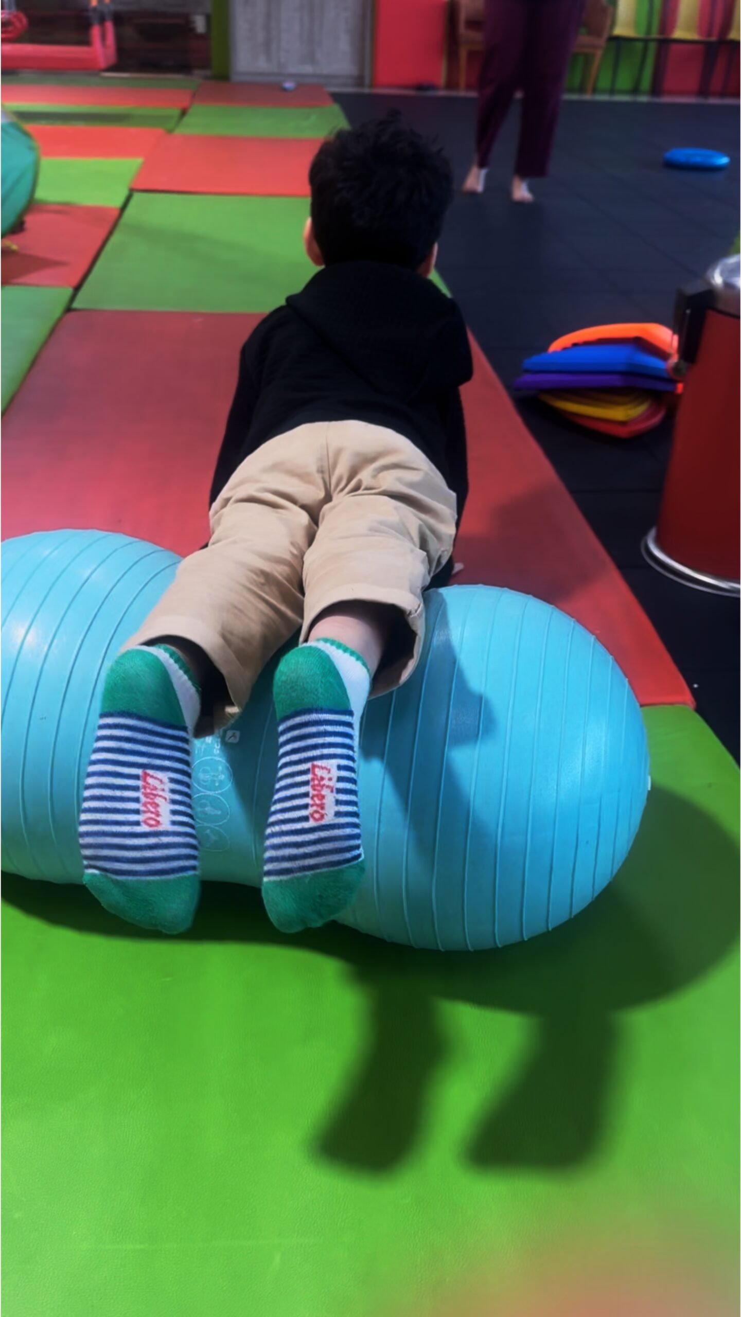 Child lying on a therapy peanut ball during a sensory integration session on colorful padded flooring.
